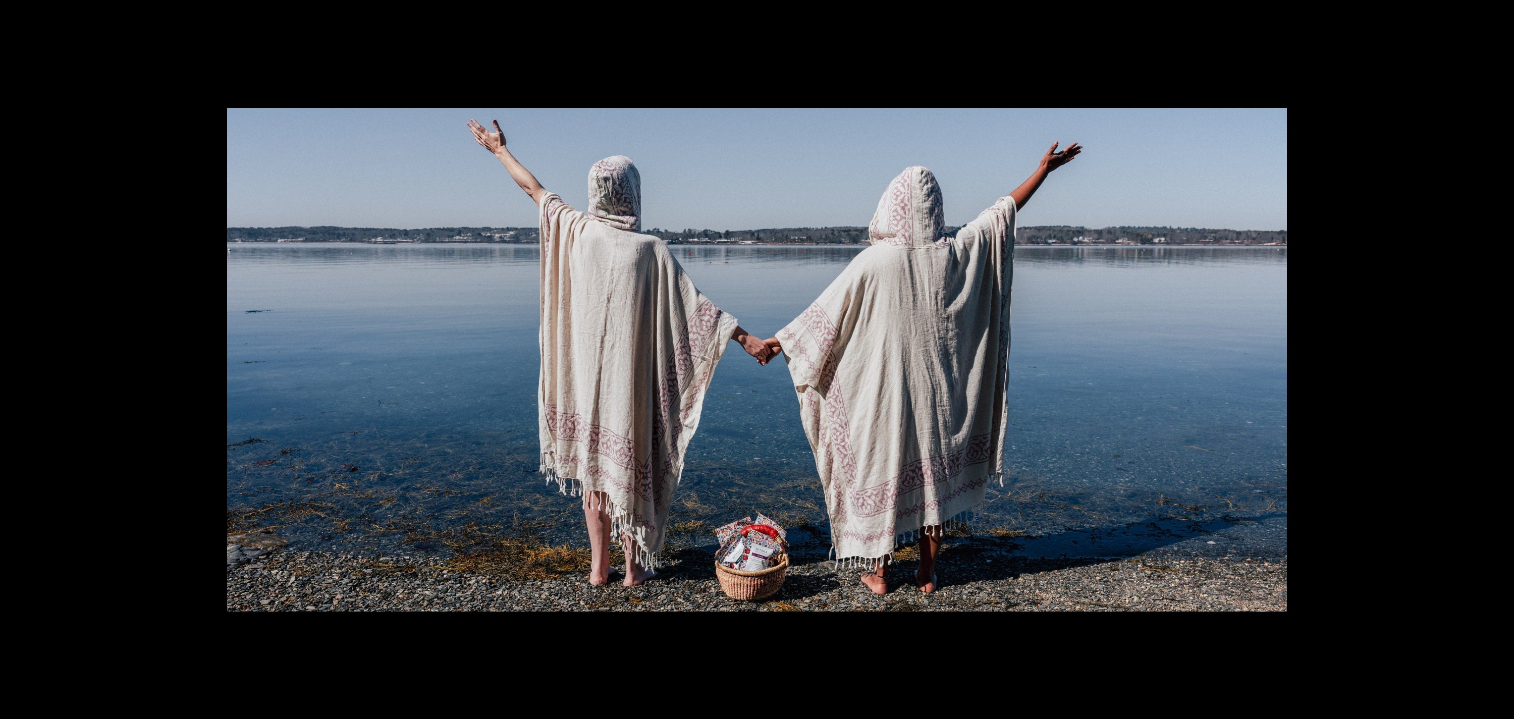 Two women in yoni steam gowns standing on a beach with arms raised, holding hands.
